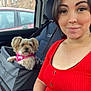 woman, dog, car_interior, pet_booster_seat, red_top, smile, seatbelt, window, pink_harness, short_hair, hand, nail_polish, steering_wheel, car_seat, brick_building, vehicle, casual, daylight, looking, portrait
