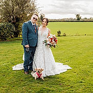 Minnie is registered to the contest to win money with this photo: wedding, bride, groom, dog, flower_bouquet, grass, outdoor, greenery, tree, dress, suit, leash, pet, smile, happy, nature, celebration, portrait, couple, cloudy_sky