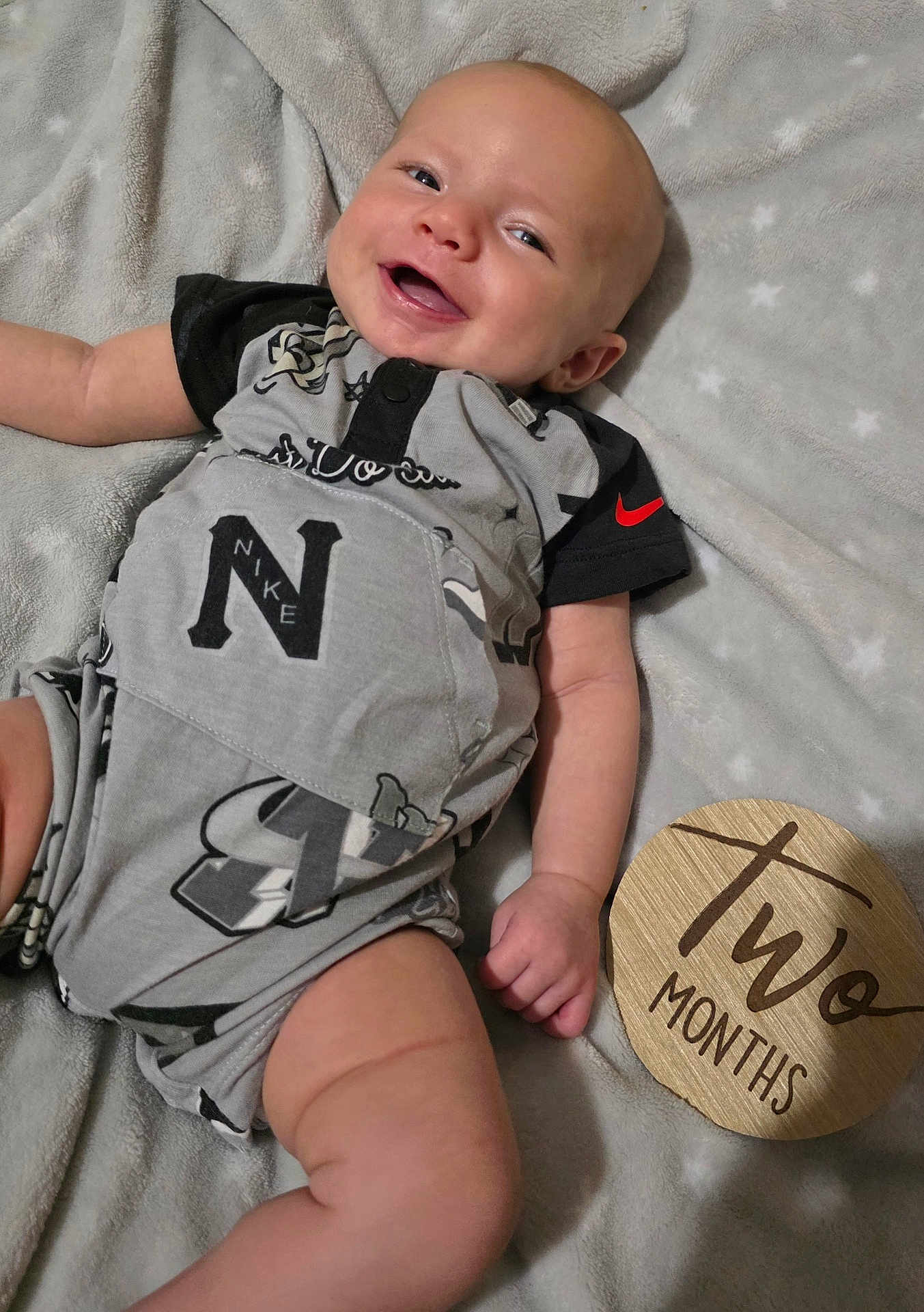 Carson joined the competition — help win amazing prizes! baby, infant, smiling, clothing, blanket, sign, wooden_sign, two_months, happy, person, face, head, skin, arm, hand, leg, foot, fabric, pattern, indoor