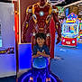 child, boy, arcade, iron_man_statue, toy, glowing, neon_lights, gaming_machine, smiling, crossed_arms, shoes, curly_hair, blue_shorts, blue_shirt, indoor, floor, background, amusement, entertainment, colorful