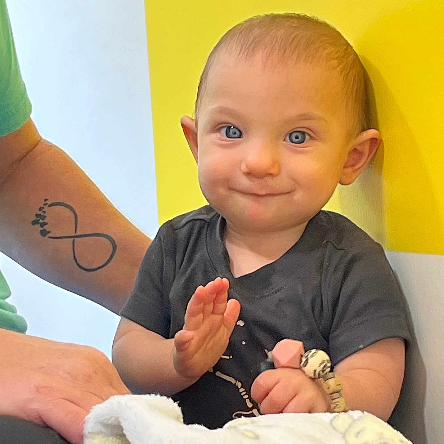 Kason is registered to the contest to win money with this photo: arm, baby, black_shirt, blanket, blue_eyes, bracelet, child, clapping, cute, happy, human, indoor, person, portrait, seated, smile, tattoo, toddler, toy, yellow_wall
