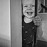 toddler, child, smiling, laughing, portrait, black_and_white, indoors, doorway, doorframe, hand, teeth, face, eyes, clothing, overalls, standing, home, interior, playful, happy