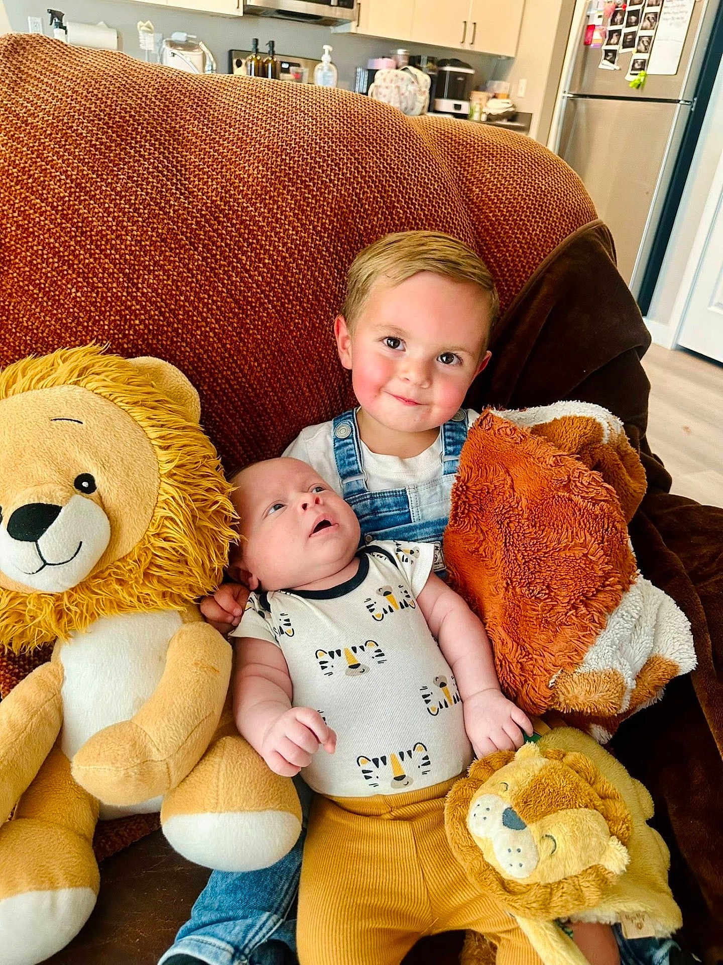 Isaiah is registered to the contest to win money with this photo: baby, child, couch, plush_toy, lion_toy, blanket, smiling, overalls, infant, living_room, furniture, cute, sibling, happy, indoor, person, toddler, clothing, face, home