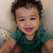 Jordan is registered to the contest to win money with this photo: baby, toddler, smiling, curly_hair, green_shirt, dinosaur_print, blanket, cushion, indoor, happy, face, person, child, portrait, cute, young, skin, eyes, mouth, sitting
