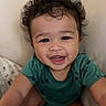 baby, toddler, smiling, curly_hair, green_shirt, dinosaur_print, blanket, cushion, indoor, happy, face, person, child, portrait, cute, young, skin, eyes, mouth, sitting