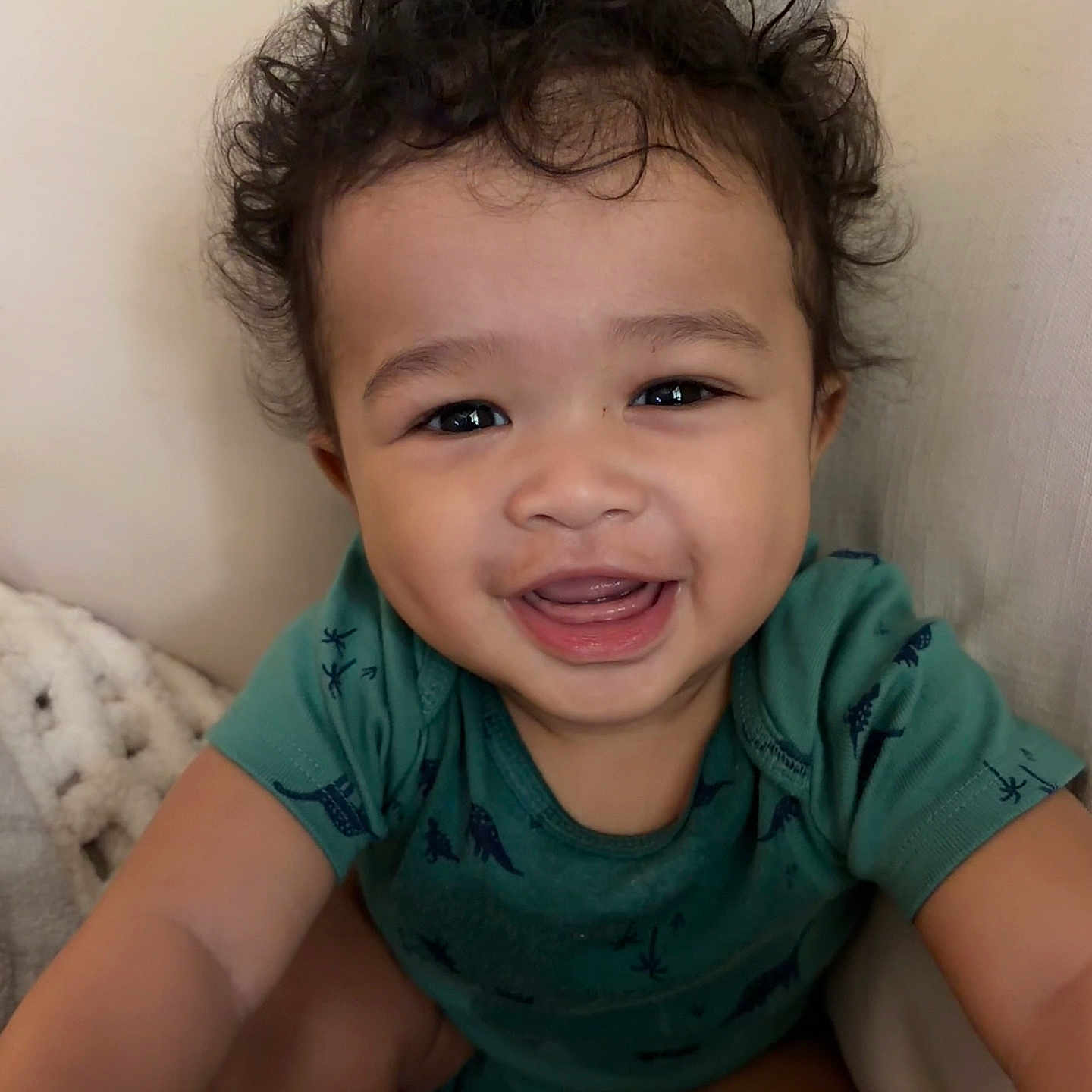 Jordan is registered to the contest to win money with this photo: baby, toddler, smiling, curly_hair, green_shirt, dinosaur_print, blanket, cushion, indoor, happy, face, person, child, portrait, cute, young, skin, eyes, mouth, sitting