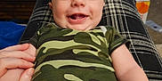 Maxwell is registered to the contest to win money with this photo: baby, bottle, camouflage_clothing, can, carpet, clothing, container, face, furniture, hand, happy, holding, indoor, infant, legs, living_room, people, person, smiling, table
