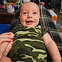 baby, bottle, camouflage_clothing, can, carpet, clothing, container, face, furniture, hand, happy, holding, indoor, infant, legs, living_room, people, person, smiling, table