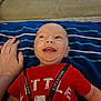 baby, blue_blanket, casual_clothing, child, cute, face, hand, happy, head, indoor, infant, lifeguard_text, lying_down, person, red_shirt, skin, smiling, striped_blanket, tongue_out, young_child