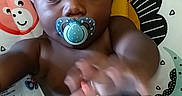 Davell is registered to the contest to win money with this photo: baby, pacifier, toy, infant, child, seat, patterned, wooden_floor, reaching, blue_toy, red_toy, face, hand, curious, indoor, playtime, young_child, black_hair, expression, closeup