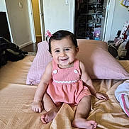 Alana is registered to the contest to win money with this photo: baby, child, smiling, pink_dress, bow, bed, pillow, indoor, person, happy, cute, hair, skin, furniture, room, blanket, sitting, foot, hand, face