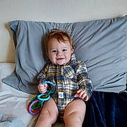 Aspen joined the competition — help win amazing prizes! baby, child, plaid_onesie, smiling, sitting, bed, pillow, blanket, toy, colorful_rings, indoor, cute, happy, person, infant, young_child, face, hand, legs, cozy