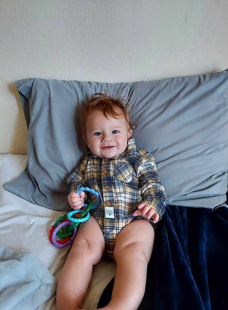 Aspen joined the competition — help win amazing prizes! baby, child, plaid_onesie, smiling, sitting, bed, pillow, blanket, toy, colorful_rings, indoor, cute, happy, person, infant, young_child, face, hand, legs, cozy