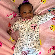 Assani is registered to the contest to win money with this photo: baby, child, smile, onesie, blanket, pink, purple_bow, milk_bottle_pattern, happy_text, lying_down, cute, infant, face, clothing, pattern, soft_texture, colorful, indoor, portrait, joyful
