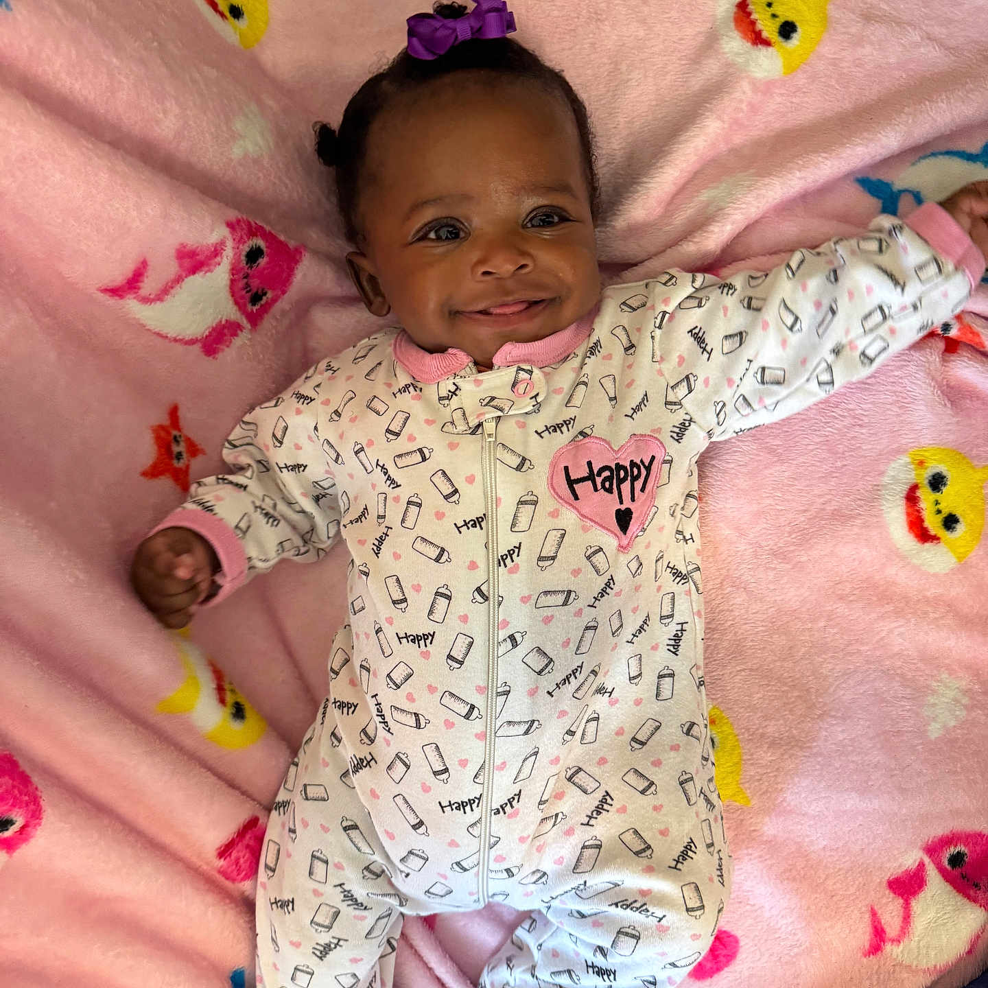 Assani is registered to the contest to win money with this photo: baby, blanket, child, clothing, colorful, cute, face, happy_text, indoor, infant, joyful, lying_down, milk_bottle_pattern, onesie, pattern, pink, portrait, purple_bow, smile, soft_texture