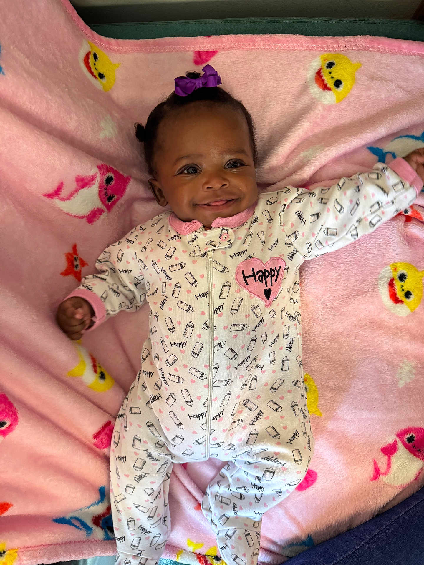 Assani is registered to the contest to win money with this photo: baby, child, smile, onesie, blanket, pink, purple_bow, milk_bottle_pattern, happy_text, lying_down, cute, infant, face, clothing, pattern, soft_texture, colorful, indoor, portrait, joyful