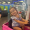 Charlie is registered to the contest to win money with this photo: child, girl, smiling, portrait, toy_car, pink_vehicle, driving, indoor, playroom, salon, ring_light, dress, hair_bows, curly_hair, seated, happy, steering_wheel, colorful, vehicle, photography