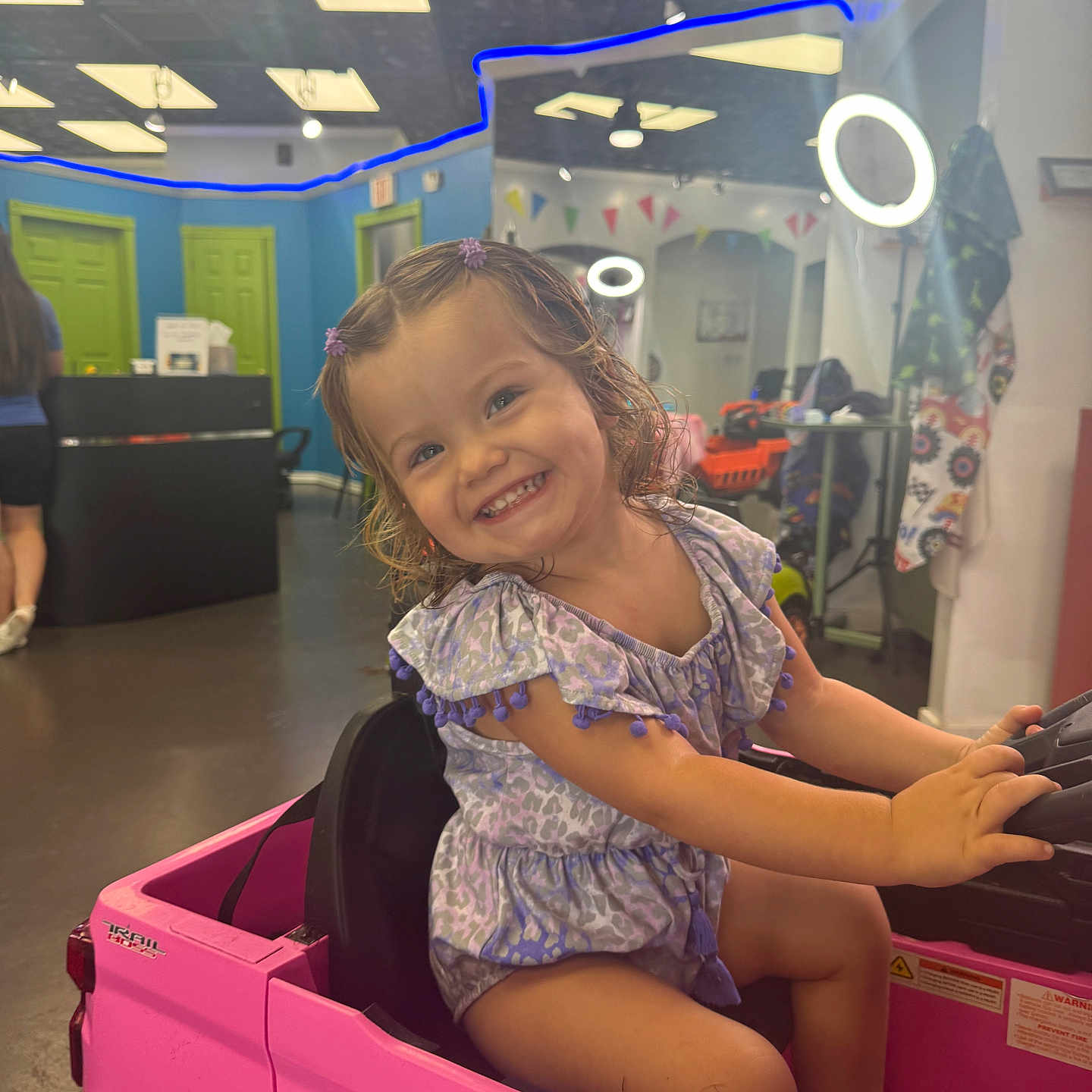 Charlie is registered to the contest to win money with this photo: child, girl, smiling, portrait, toy_car, pink_vehicle, driving, indoor, playroom, salon, ring_light, dress, hair_bows, curly_hair, seated, happy, steering_wheel, colorful, vehicle, photography