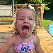 Charlie is registered to the contest to win money with this photo: child, toddler, girl, portrait, smiling, tongue_out, outdoor, summer, sunlight, wooden_deck, backyard, garden, poolside, playful, happy, blonde_hair, pink_clothing, swimsuit, close_up, face