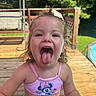 child, toddler, girl, portrait, smiling, tongue_out, outdoor, summer, sunlight, wooden_deck, backyard, garden, poolside, playful, happy, blonde_hair, pink_clothing, swimsuit, close_up, face