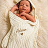 baby, newborn, infant, wrapped, towel, hooded_towel, blanket, soft_texture, cream_color, embroidered_name, heart_symbol, lying_down, curious_expression, small_hands, face, skin, cozy, fluffy_rug, portrait, close_up