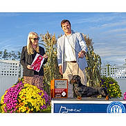 Sebastian is registered to the contest to win money with this photo: dog, dachshund, award, ribbon, podium, american_kennel_club, person, man, woman, flowers, cornstalks, outdoor, sunny, smiling, blue_sky, event, competition, proud, leash, clothing