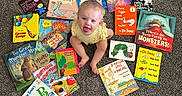 Bexley joined the competition — help win amazing prizes! baby, child, infant, sitting, smiling, open_mouth, portrait, books, childrens_books, storybook, reading, carpet, indoor, colorful, yellow_clothing, overhead_view, playful, happy, education, book_collection