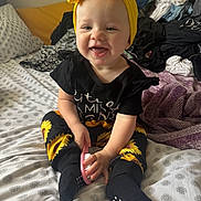 Bexley is registered to the contest to win money with this photo: baby, child, infant, smiling, happy, portrait, indoor, bedroom, bed, bedding, clothing, laundry, yellow_bow, headband, sunflower, black_outfit, hairbrush, sitting, patterned_blanket, close_up