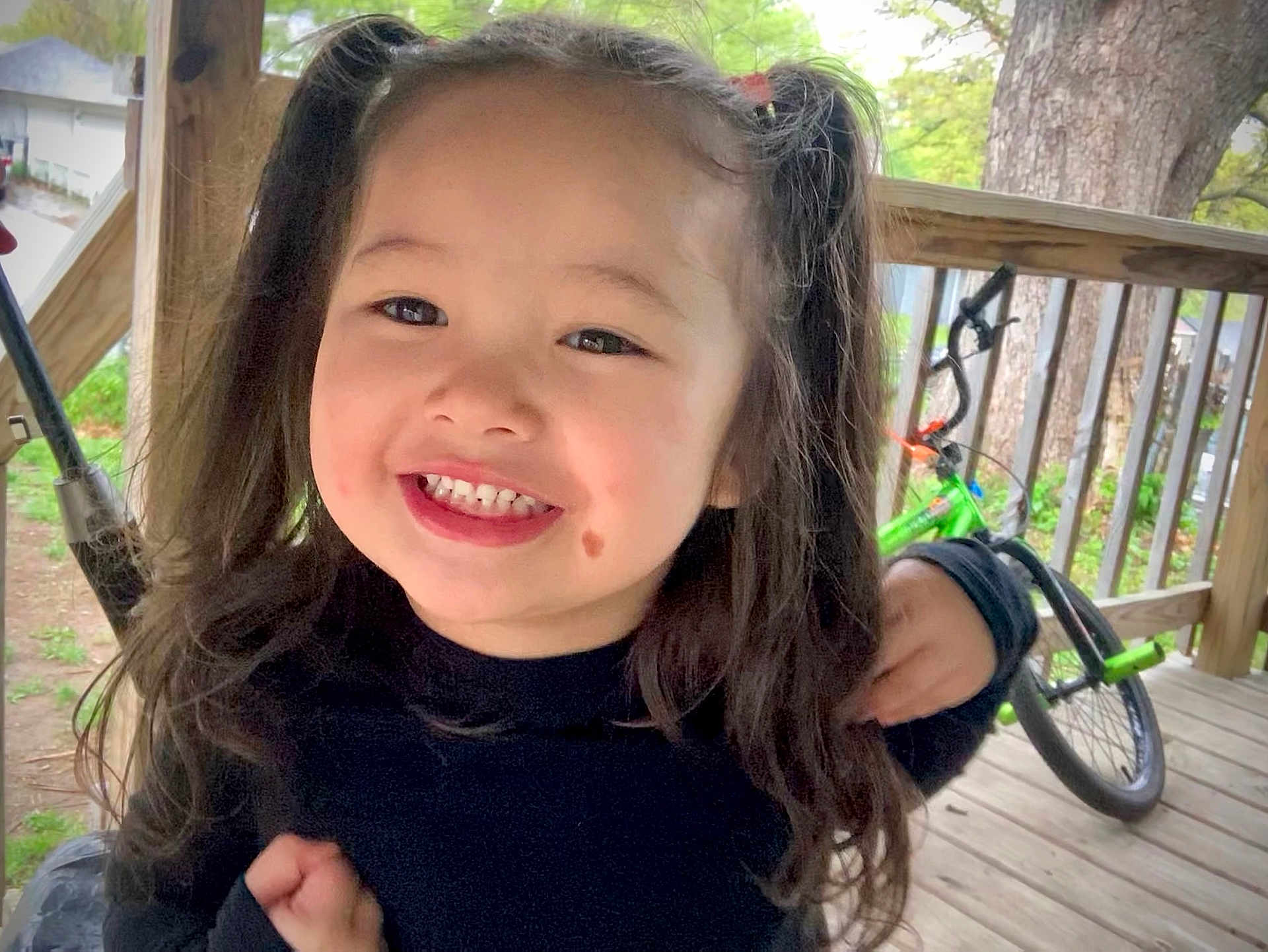Jaimelynn joined the competition — help win amazing prizes! child, smiling, pigtails, outdoor, porch, wooden_floor, bicycle, green_bike, happy, person, face, hair, casual, daylight, nature, tree, railing, clothing, cute, young
