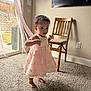 Elara joined the competition — help win amazing prizes! toddler, child, pink_dress, ribbon, indoor, carpet, barefoot, curly_hair, wooden_chair, wall, power_outlet, window, daylight, poster, curious, standing, home_interior, person, young_child, exploring