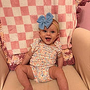 Cassidy joined the competition — help win amazing prizes! baby, child, smile, blue_bow, floral_outfit, blanket, checkered_pattern, pink, beige_chair, happy, cute, indoor, infant, headband, seated, legs, arms, face, portrait, cozy