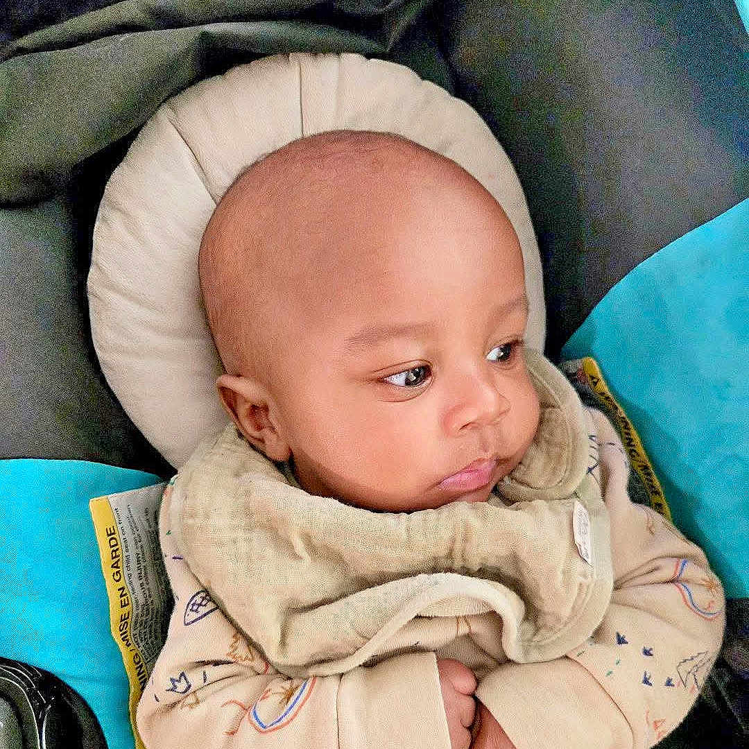 Kai is registered to the contest to win money with this photo: baby, infant, child, portrait, face, eyes, bald, bundled, blanket, clothing, sitting, car_seat, cushion, indoor, close_up, looking_away, cozy, text_overlay, emoji, family_photo
