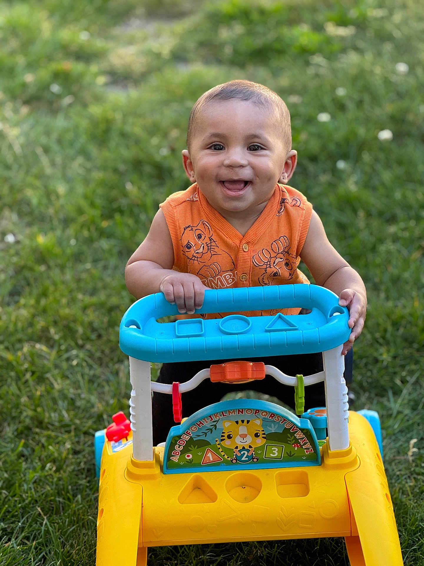 Zahkai is registered to the contest to win money with this photo: toddler, child, smiling, outdoor, grass, toy, walker, yellow, blue, orange_clothing, happy, cute, baby, play, summer, green, park, fun, person, portrait