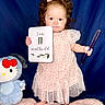baby, child, infant, girl, pink_dress, barefoot, holding_card, milestone, hello_kitty, plush_toy, blue_background, soft_blanket, sparkly_wand, curly_hair, bow, standing, portrait, indoors, cute, celebration
