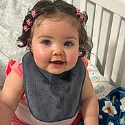 Bridget is registered to the contest to win money with this photo: baby, child, crib, bib, pink_clothing, curly_hair, hair_clips, blue_eyes, blanket, smiling, indoors, baby_girl, cute, sitting, infant, face, hand, soft_lighting, home, portrait