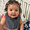 baby, child, crib, bib, pink_clothing, curly_hair, hair_clips, blue_eyes, blanket, smiling, indoors, baby_girl, cute, sitting, infant, face, hand, soft_lighting, home, portrait