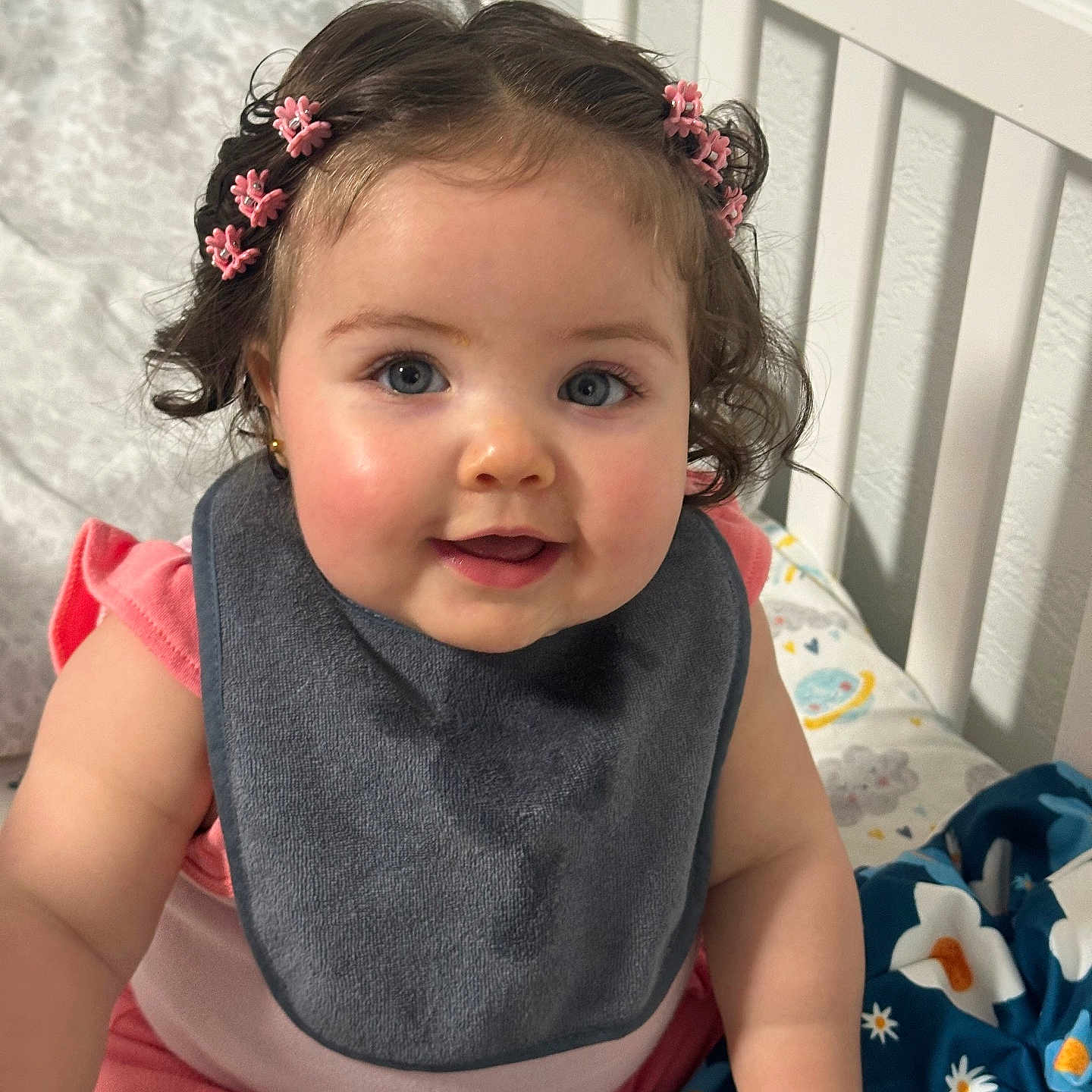 Bridget is registered to the contest to win money with this photo: baby, child, crib, bib, pink_clothing, curly_hair, hair_clips, blue_eyes, blanket, smiling, indoors, baby_girl, cute, sitting, infant, face, hand, soft_lighting, home, portrait