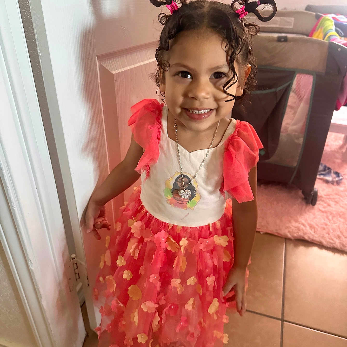Aya is registered to the contest to win money with this photo: carpet, child, curly_hair, door, dress, floor, flowers, girl, hair, happy, home, indoor, jewelry, necklace, person, pigtails, pink, playful, smile, tile