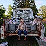 child, toddler, smiling, sitting, vintage_truck, rustic, wooden_bench, flowers, flower_buckets, outdoor, greenery, grass, tree, sky, rust, denim_jeans, checkered_shirt, boots, watering_can, garden