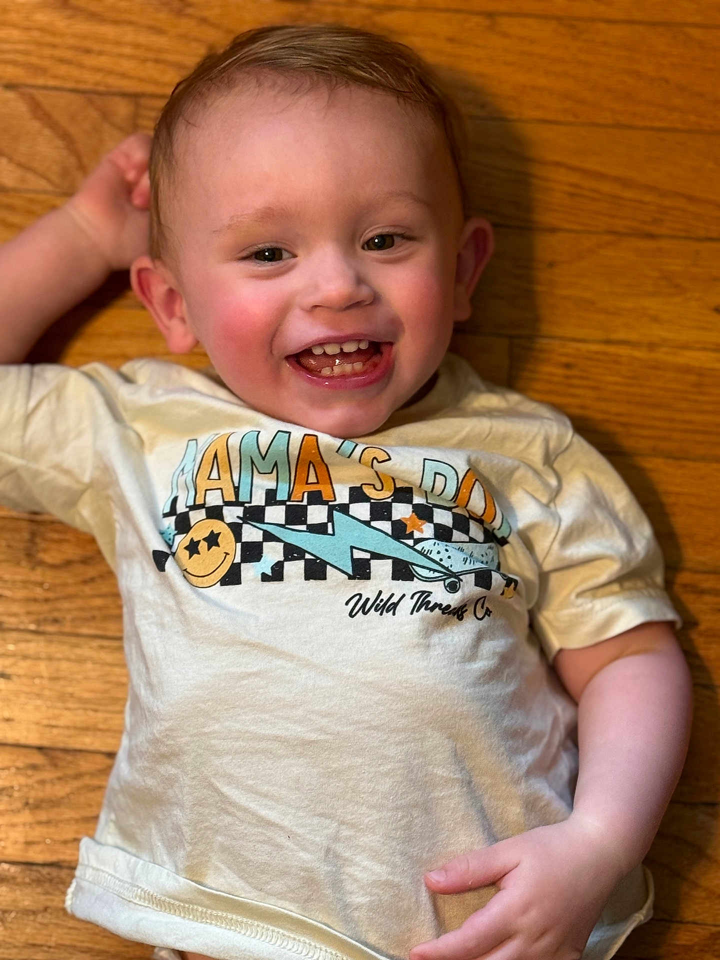 Jackson is registered to the contest to win money with this photo: toddler, child, smiling, happy, baby_teeth, tshirt, wooden_floor, laying_down, cute, rosy_cheeks, casual_clothing, indoor, portrait, young_child, playful, graphic_tee, expression, person, head, hand