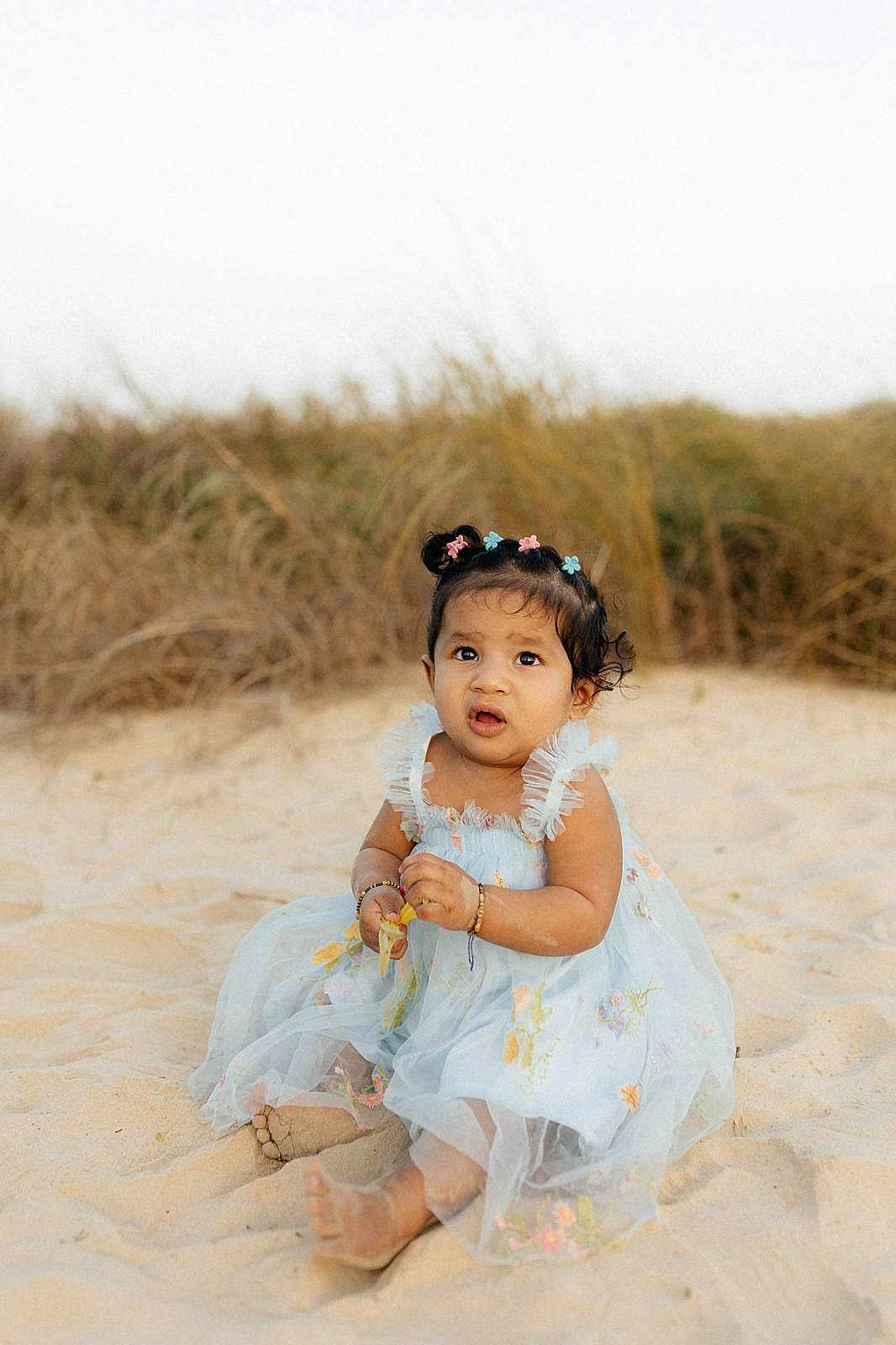 Aria joined the competition — help win amazing prizes! child, toddler, baby, dress, floral_dress, beach, sand, dunes, grass, sitting, barefoot, hair_bows, bracelets, curious_expression, portrait, outdoor, candid, cute, sunshine, hands