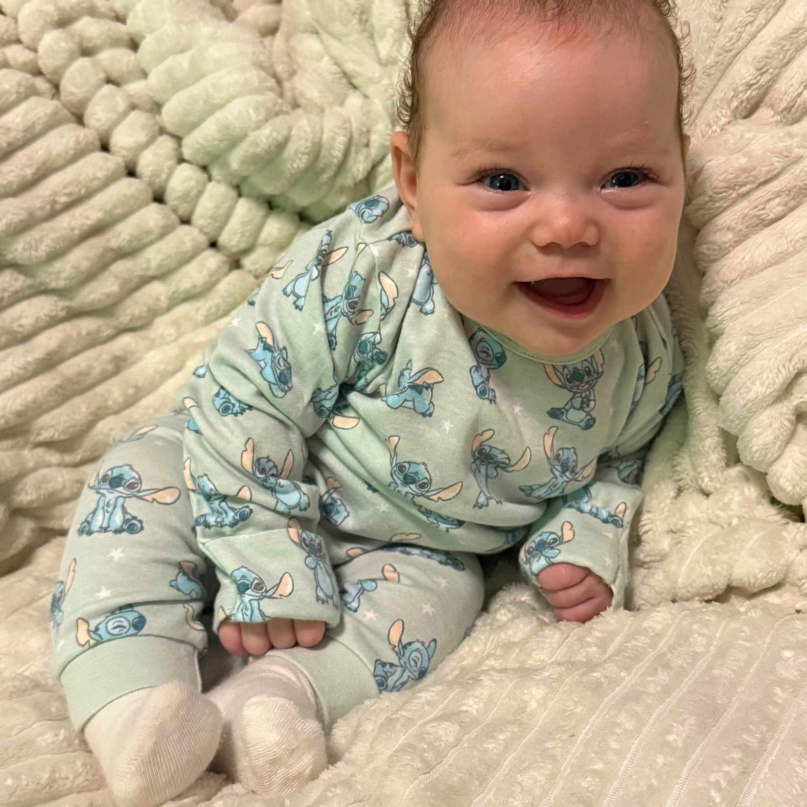 Millie-Rose is registered to the contest to win money with this photo: baby, child, cozy, curly_hair, cute, face, footwear, happy, indoors, infant, light_skin, pajamas, patterned_clothing, plush_blanket, portrait, sitting, small_hands, smiling, soft_texture, stitch