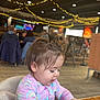 baby, background, casual_clothing, child, curly_hair, daylight, dinosaur_onesie, focused, hair, high_chair, indoor, lights, people, public_space, restaurant, seated, string_lights, table, toddler, wooden_table