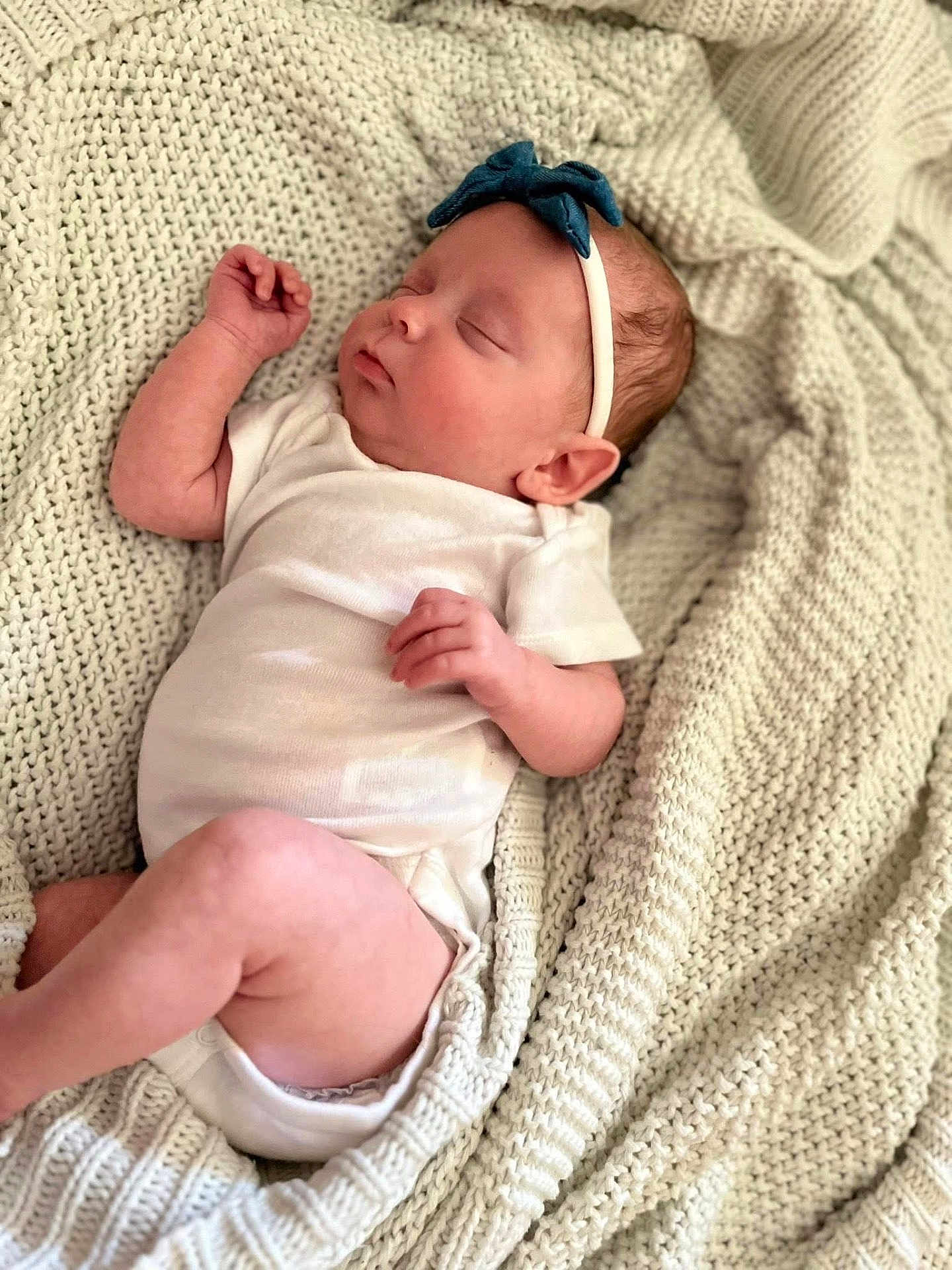 Emery is registered to the contest to win money with this photo: baby, newborn, sleeping, onesie, headband, blue_bow, blanket, knitted_blanket, cozy, infant, peaceful, cute, soft, portrait, indoor, person, skin, hand, leg, closeup