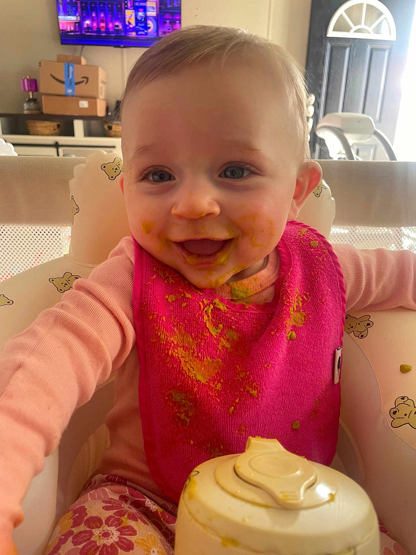 Kasey is registered to the contest to win money with this photo: baby, infant, toddler, highchair, bib, bib_stained, baby_food, messy, smile, face, eyes, clothing, pink, feeding_bottle, food_container, indoor, living_room, television, door, happy