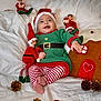 baby, celebration, child, christmas_elf, cozy, cute, feet, festive, gingerbread_house, green_outfit, happy, holiday_decor, indoors, pinecone, plush_toy, red_hat, smiling, striped_pants, toy, white_blanket