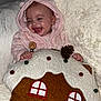 baby, child, hood, pink_clothing, smiling, laughing, pillow, gingerbread_house, pom_poms, pinecone, blanket, soft_texture, cozy, indoors, cute, happy, infant, warm, clothing, portrait