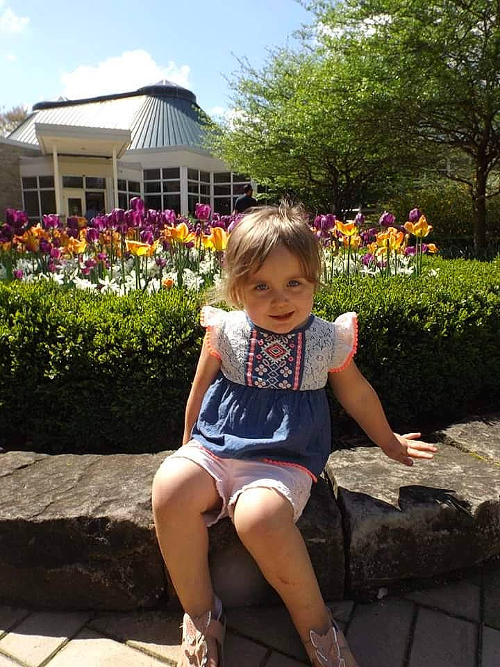 Emma is registered to the contest to win money with this photo: botany, child, flower, footwear, fun, garden, grass, leg, leisure, person, pink, plant, shoe, sitting, smile, spring, summer, thigh, toddler, tourism