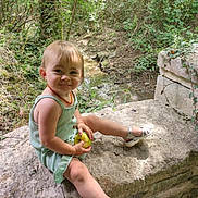 Apolline a rejoint le concours — aidez-le/la à gagner de superbes lots ! toddler, child, outdoor, forest, nature, apple, sandals, greenery, stone, smile, happy, sunlight, trees, dress, person, sitting, playful, young_child, daylight, cute