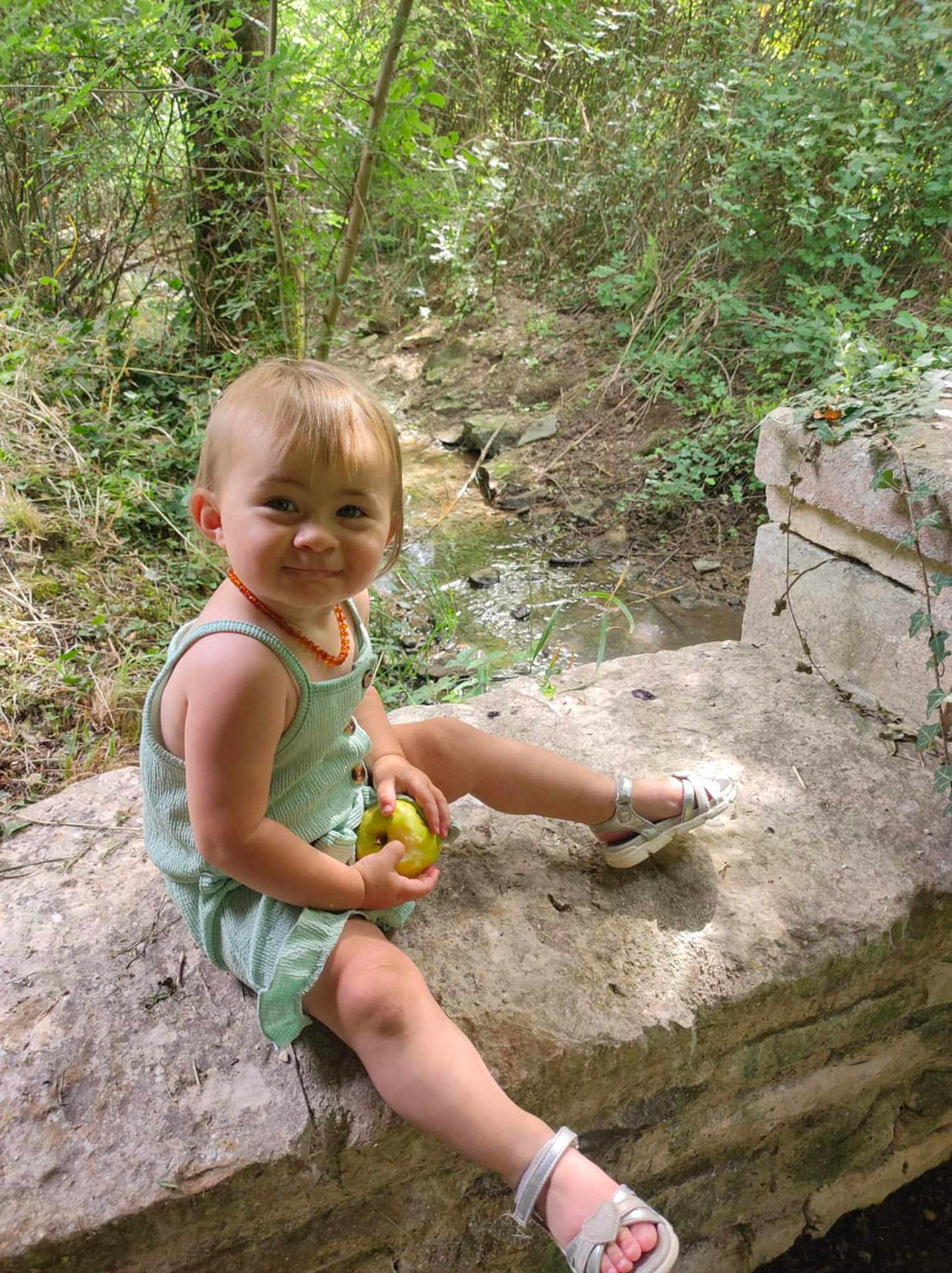 Apolline a rejoint le concours — aidez-le/la à gagner de superbes lots ! toddler, child, outdoor, forest, nature, apple, sandals, greenery, stone, smile, happy, sunlight, trees, dress, person, sitting, playful, young_child, daylight, cute